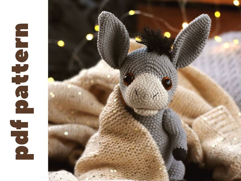 May include: A gray crocheted donkey toy with black yarn for the mane and tail. The donkey is sitting on a beige knitted blanket with gold glitter.