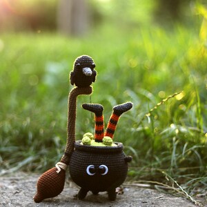 May include: A crocheted witch with black and orange striped legs sits in a black cauldron with green goo inside. A black crocheted crow sits on top of the witch's broom.