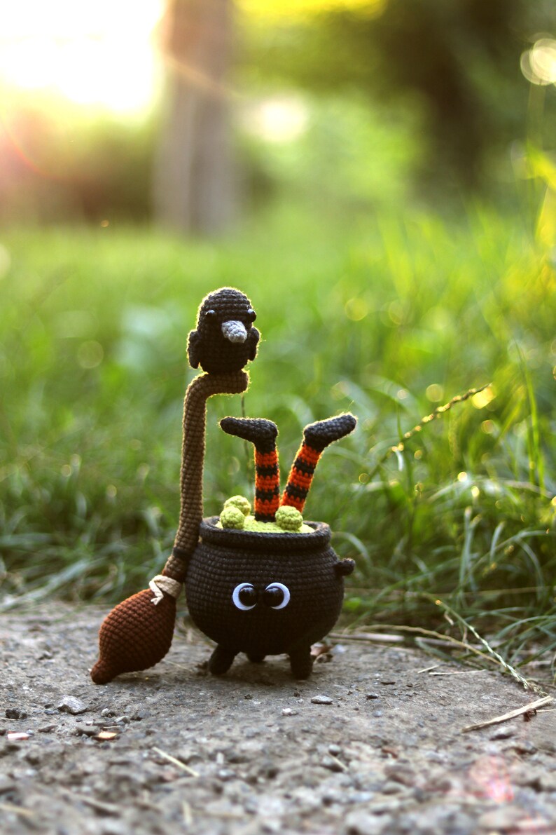May include: A crocheted witch with black and orange striped legs sits in a black cauldron with green goo inside. A black crocheted crow sits on top of the witch's broom.