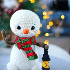 May include: A white crocheted snowman with a red and green striped scarf and a black crocheted lantern. The snowman has a carrot nose and black eyes.