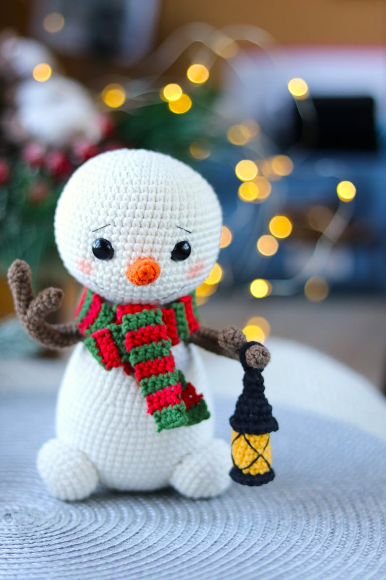May include: A white crocheted snowman with a red and green striped scarf and a black crocheted lantern. The snowman has a carrot nose and black eyes.