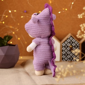 May include: A purple crocheted stuffed animal shaped like a dinosaur. The dinosaur is standing on a light brown surface.