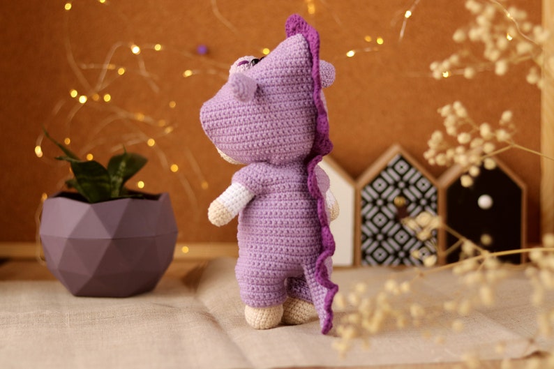 May include: A purple crocheted stuffed animal shaped like a dinosaur. The dinosaur is standing on a light brown surface.