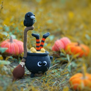 May include: A black crocheted cauldron with a witch's legs sticking out of the top. A black crocheted raven sits on a brown stick attached to the cauldron. The cauldron is surrounded by orange pumpkins.