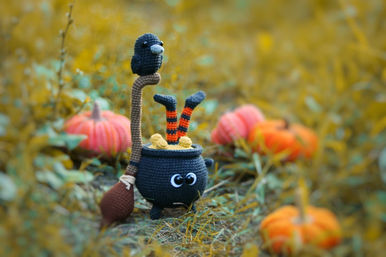 May include: A black crocheted cauldron with a witch's legs sticking out of the top. A black crocheted raven sits on a brown stick attached to the cauldron. The cauldron is surrounded by orange pumpkins.