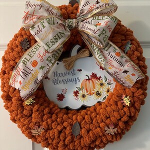 May include: A brown yarn wreath decorated with fall-themed ribbon and a pumpkin sign that reads "Harvest Blessings". The wreath is adorned with small, brown leaves.