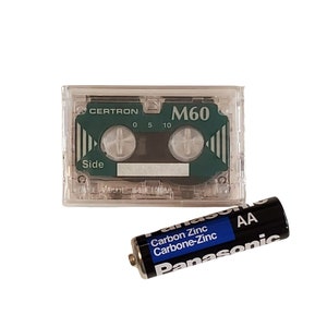 May include: A clear plastic cassette tape labeled "Certron M60" with a green label and the words "Side" and "Made in China". A black AA battery with blue and white text that reads "Panasonic Carbon Zinc Carbone-Zinc" is also in the image.