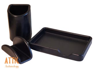 May include: A black desk organizer set featuring a pen holder, a small tray, and a curved phone or card holder. The set is made of a solid, matte black material. The text "ATTIC Archaeology" is visible.