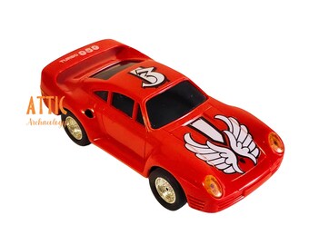 May include: A red toy car with the text "TURBO 059" on the spoiler. The car has a white and black graphic on the hood, the number "3" on the roof, and gold-colored wheels. The car is a vintage toy.