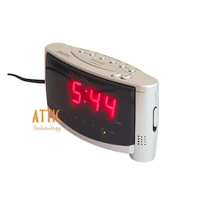 May include: Silver digital alarm clock displaying 5:44 in bright red numerals. The clock has several buttons on top and a curved black display screen. The brand name is visible on the top of the clock. The clock is plugged into a black power cord.