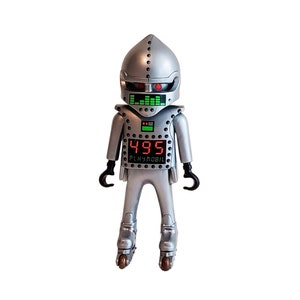 May include: A silver Playmobil robot figure with a helmet, roller skates, and black claw-like hands. The robot has a digital display showing the number "495" in red and a green equalizer. The word "PLAYMOBIL" is printed below the number.