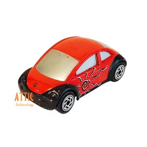 May include: A small, red toy car with black accents and silver roof. The car features a sun-like design on the side and a Volkswagen logo on the front. The wheels are black with silver rims. The car is a collectible item.