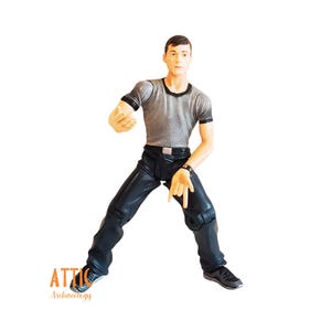 May include: A figurine of a man in a gray t-shirt with black trim and black leather-look pants. The figure is posed with one arm extended and the other hand pointing down. The figure is wearing a black watch and black shoes.