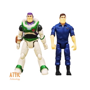 Peut inclure: Deux figurines du film Toy Story. L'une est Buzz l'&Eacute;clair dans une combinaison spatiale blanche, verte et violette. L'autre est un homme en uniforme bleu. Les figurines sont sur fond blanc. Le logo Attic Archaeology est visible.