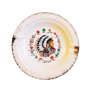 May include: A white ceramic ashtray with a Native American chief design. The design includes various symbols and text, including "Butterfly", "4 Ages", "Bear Track", "Crossed Arrows", "Eagle Feathers", "Thunderbird", "Paths", "Tepee", "Candle", "Back", "Running Water", "Arrow", "Arrow Head", "Bear Track", "Cactus", "Horse", and "Sun Symbols".