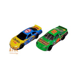 May include: Two toy race cars. One is yellow and blue with the number 43 and the Cheerios logo. The other is green with the number 18 and the Interstate Batteries logo. Both have black wheels.