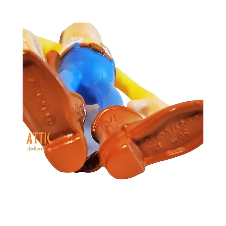 May include: A close-up of a toy figure, featuring brown boots, blue pants, and a yellow shirt. The figure's feet are in focus, with details of the soles visible. The figure is on a white background.