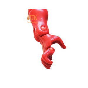 Puede incluir: Un brazo rojo brillante, articulado, posiblemente un elemento decorativo o parte de una escultura m&aacute;s grande. El brazo est&aacute; doblado en el codo y la mu&ntilde;eca, con la mano parcialmente abierta. El logotipo "ATTIC Archeology" es visible.