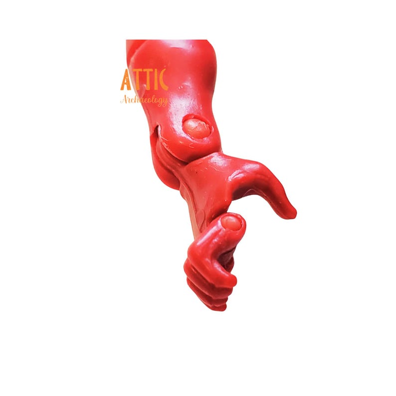 Puede incluir: Un brazo rojo brillante, articulado, posiblemente un elemento decorativo o parte de una escultura m&aacute;s grande. El brazo est&aacute; doblado en el codo y la mu&ntilde;eca, con la mano parcialmente abierta. El logotipo "ATTIC Archeology" es visible.