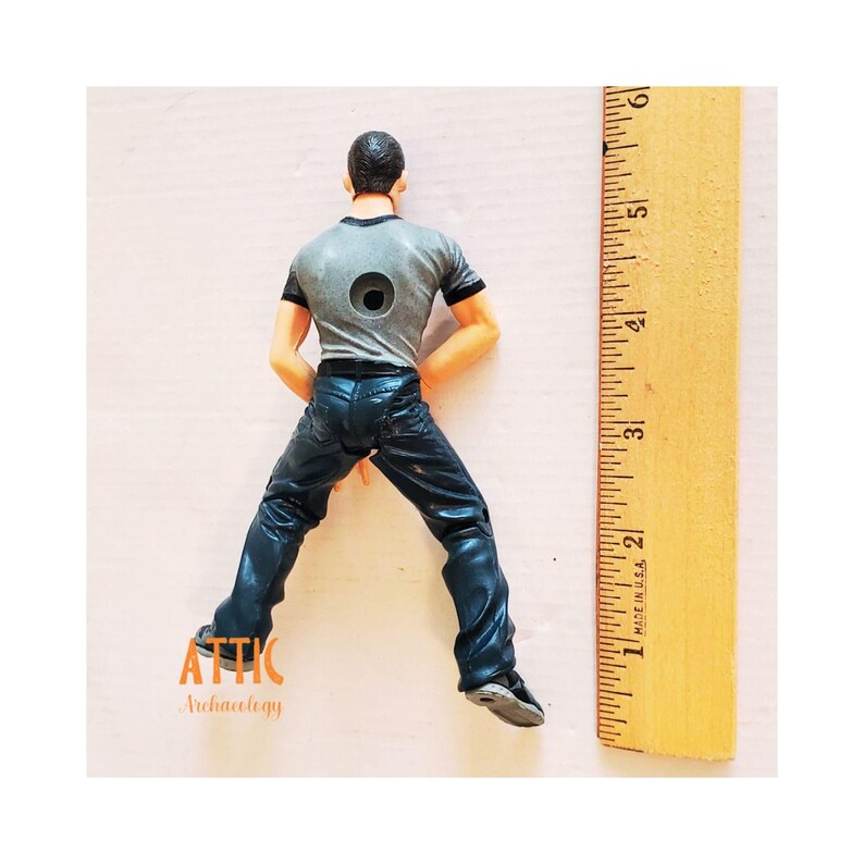 May include: A figurine of a person wearing a gray t-shirt and dark blue jeans. The figurine is posed with legs spread and arms at the sides. A wooden ruler is next to the figurine, showing measurements in inches.