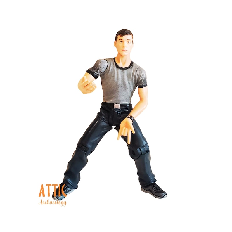 May include: A figurine of a man in a gray t-shirt with black trim and black leather-look pants. The figure is posed with one arm extended and the other hand pointing down. The figure is wearing a black watch and black shoes.