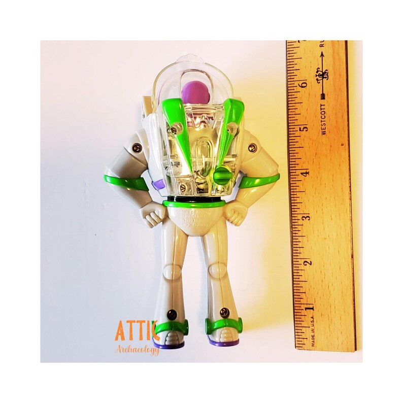 May include: A white and green plastic Buzz Lightyear action figure with a clear plastic helmet and a purple visor. The figure is standing with its arms outstretched and is approximately 5 inches tall.
