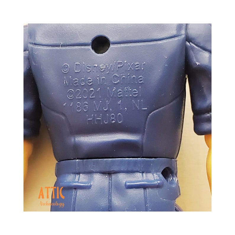 Peut inclure: Gros plan sur le dos d'une figurine en plastique bleu. Le texte sur la figurine indique "&copy; Disney/Pixar Made in China &copy;2021 Mattel 1186 MJ, 1, NL HHJ80". Le mot "ATTIC" est &eacute;galement visible.