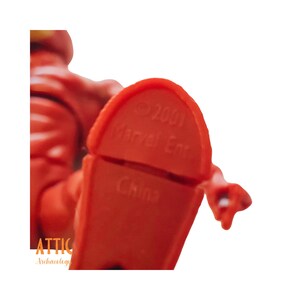 Puede incluir: Primer plano del pie de una figura de acci&oacute;n de pl&aacute;stico rojo. El pie est&aacute; estampado con "&copy; 2001 Marvel Ent" y "China". La figura es parcialmente visible, de color rojo brillante.
