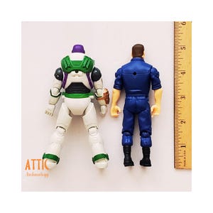 Peut inclure: Deux figurines, Buzz l'&Eacute;clair et un personnage en uniforme, sont pr&eacute;sent&eacute;es de dos. Buzz l'&Eacute;clair est blanc, vert et violet. Le personnage en uniforme est bleu. Une r&egrave;gle montre des mesures en centim&egrave;tres.