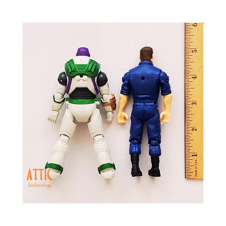 Peut inclure: Deux figurines, Buzz l'&Eacute;clair et un personnage en uniforme, sont pr&eacute;sent&eacute;es de dos. Buzz l'&Eacute;clair est blanc, vert et violet. Le personnage en uniforme est bleu. Une r&egrave;gle montre des mesures en centim&egrave;tres.