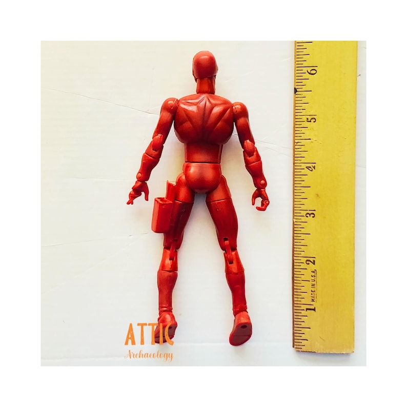 Puede incluir: Una figura de acci&oacute;n roja con un cintur&oacute;n de herramientas, junto a una regla amarilla. La figura mide aproximadamente 15 cm de altura. La figura es de color rojo s&oacute;lido y tiene articulaciones. La regla est&aacute; marcada en pulgadas.