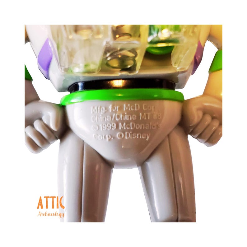 May include: A white plastic Buzz Lightyear toy with a green belt and a black belt buckle. The toy has a green and purple space ranger suit. The toy's feet are not visible in the image. The text on the toy reads: "Mig. for McD Corp China/Chine MT 88 1999 McDonald's Corp. Disney"