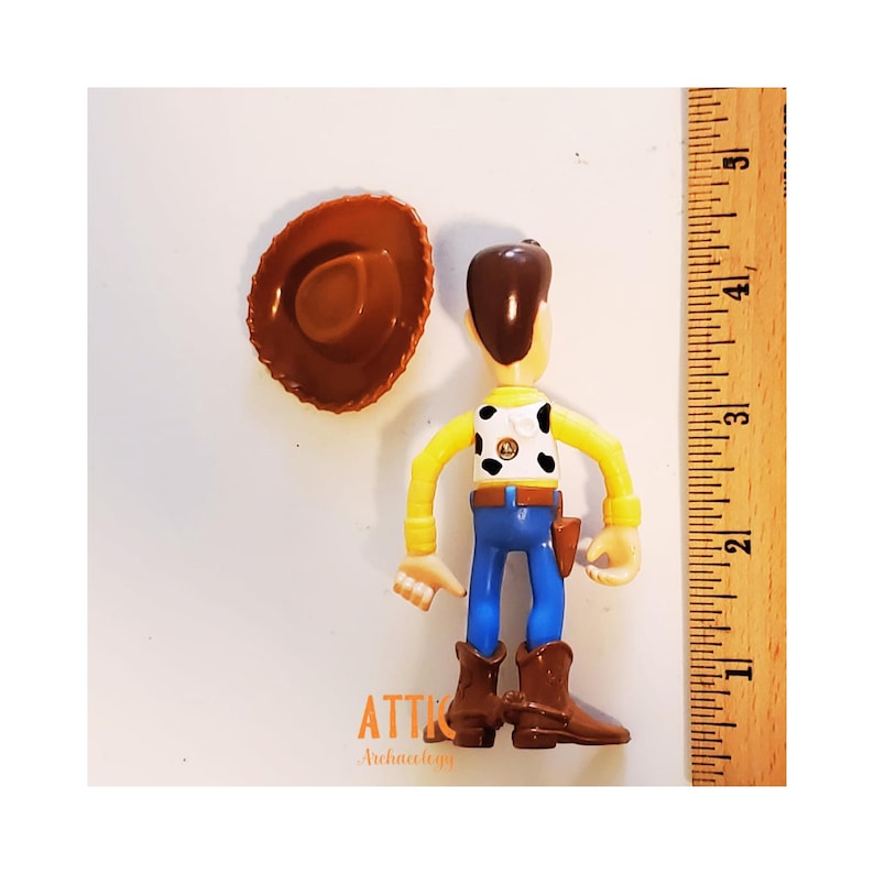 May include: A Woody action figure with a brown cowboy hat. The figure has a yellow shirt, blue pants, and brown boots. The figure is approximately 4 inches tall.