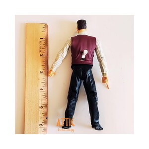 May include: A vintage action figure of a man wearing a burgundy vest, striped shirt, and black pants. The figure is approximately 6 inches tall, measured with a wooden ruler. The figure's back has a number 7 cutout. The figure is from Attic Archaeology.