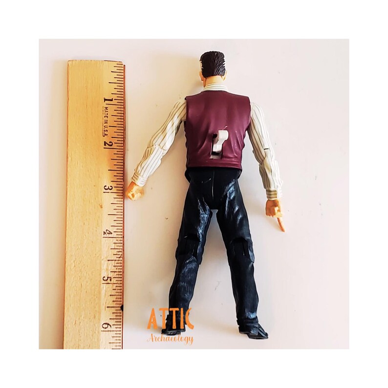 May include: A vintage action figure of a man wearing a burgundy vest, striped shirt, and black pants. The figure is approximately 6 inches tall, measured with a wooden ruler. The figure's back has a number 7 cutout. The figure is from Attic Archaeology.