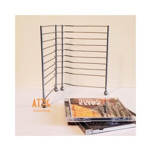 May include: A silver-toned metal wire rack designed for storing compact discs, featuring an L-shaped frame with multiple horizontal slots and rounded feet. Two compact disc cases are positioned in front of the rack, one showing the Jimmy Cliff album.