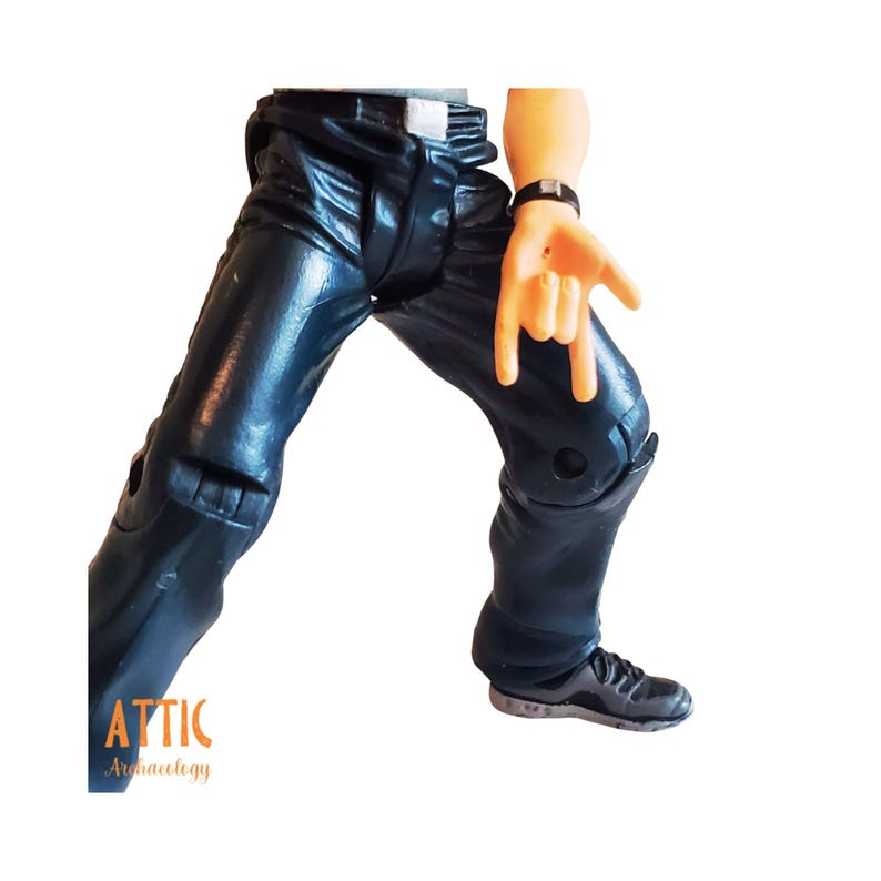 May include: A close-up of a toy figure's legs and arm. The figure wears black pants and a black watch. The figure's hand is making a rock and roll hand gesture. The word "ATTIC" is visible in the lower left corner.