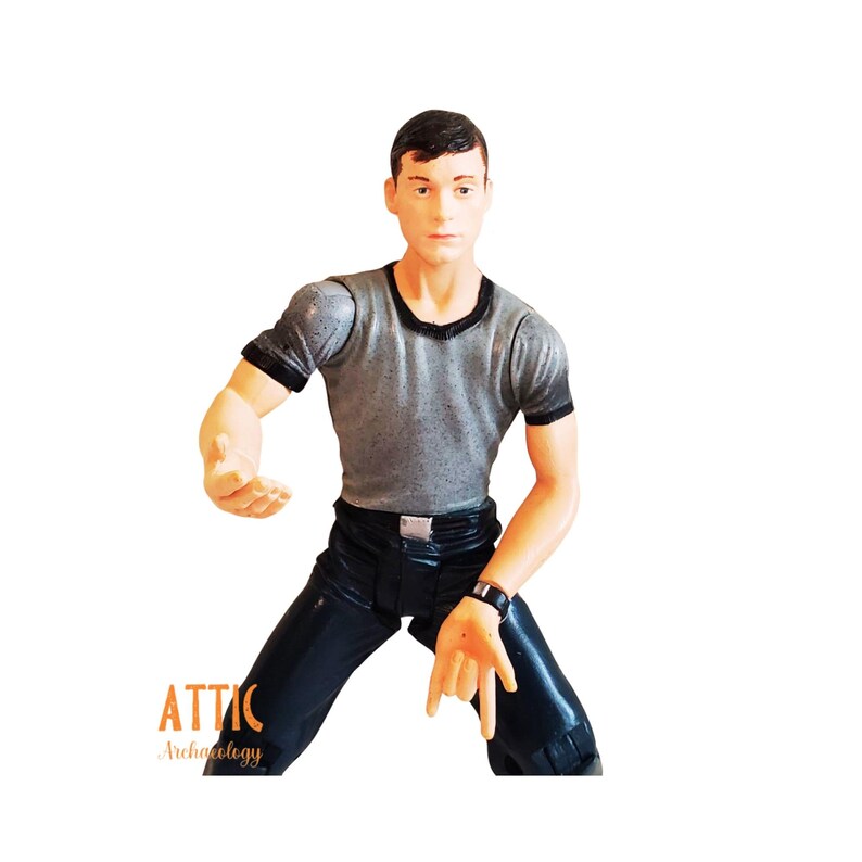 May include: A plastic action figure of a man in a gray t-shirt and black pants. The figure is posed with one arm extended and the other bent. A black watch is on the figure's wrist. The figure is against a white background.