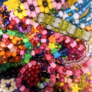 Op de afbeelding: Een close-up van een stapel kleurrijke kralenarmbanden. De armbanden zijn gemaakt met verschillende kleuren plastic kralen, waaronder rood, blauw, groen, geel en roze. Sommige armbanden hebben stervormige bedels.