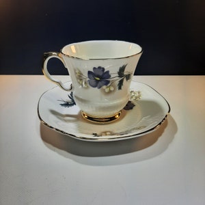 May include: A white teacup and saucer with a floral design featuring blue and white flowers. The cup has a gold rim and a gold band around the base.