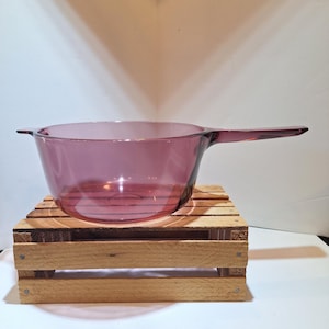 May include: A translucent purple glass saucepan featuring a round basin, a long handle extending from one side, and a small tab handle on the opposite side. The cookware has a smooth, polished surface and a deep, uniform violet hue throughout.