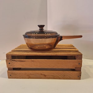 May include: A vintage amber glass saucepan with a lid and a long handle. The cookware is displayed on a rustic wooden crate, showcasing its classic design. The glass has a warm, transparent hue.
