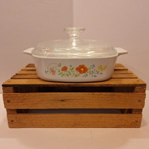 May include: White casserole dish with a glass lid. The dish has a floral design with red, orange, yellow, blue, and green flowers. The dish is sitting on a wooden crate.
