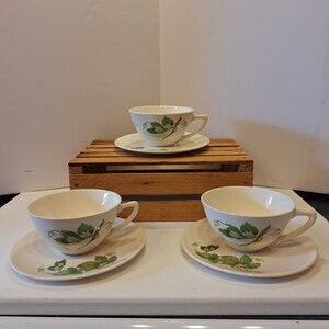 May include: Three white teacups and saucers with a green leaf design. The cups and saucers are on a wooden crate.