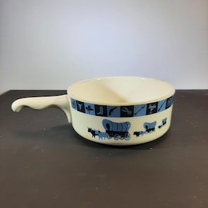 May include: Cream-colored ceramic bowl with a handle, featuring a blue and black western-themed design. The bowl has a rim with alternating blue squares with western icons and a repeating pattern of covered wagons.