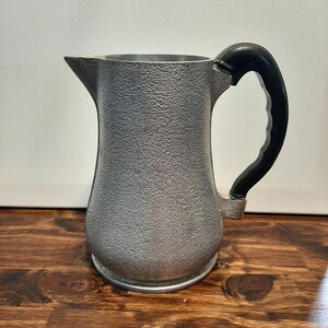 May include: A silver aluminum pitcher with a black plastic handle. The pitcher has a textured surface and a rounded body.