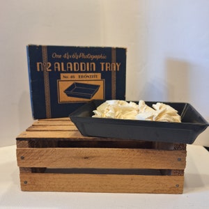 May include: A vintage black plastic photographic tray labeled "No. 46 Ebonite" and "No. 2 Aladdin Tray" in a blue box. The tray is sitting on a wooden crate.