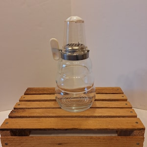 May include: A clear glass bottle with a textured surface and a silver lid. The bottle has a white plastic top with a measuring scale. The bottle is sitting on a wooden surface.
