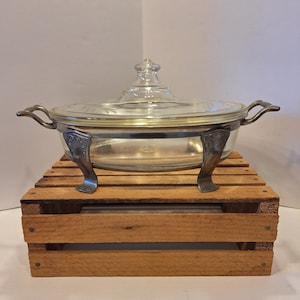 May include: A vintage glass serving dish with a silver-tone base and lid. The dish has a round shape and sits on four decorative feet. The dish is on a wooden crate.