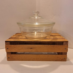 May include: A clear glass bowl with a matching lid, resting on a small wooden crate. The bowl has a rounded shape with a lid that has a small knob. The wooden crate is made of light brown wood.